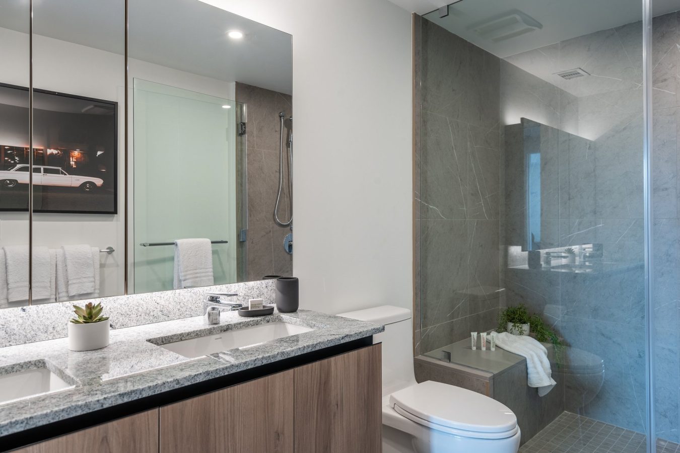 Modern bathroom with a large mirror, double sinks, wood cabinetry, a white toilet, and a glass-enclosed shower. Towels, candles, and a small plant add decorative touches to the gray and white space.