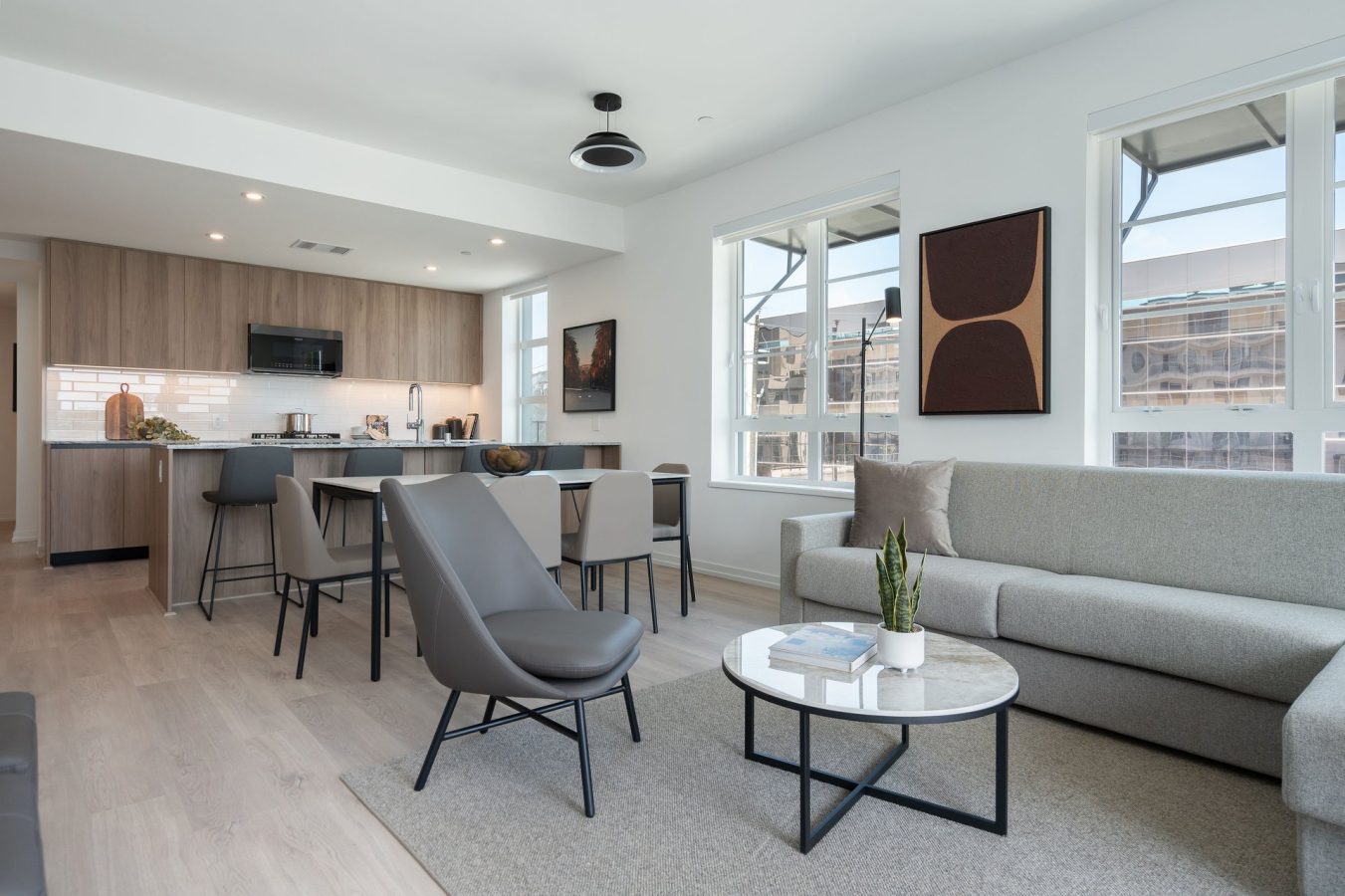 Modern open-plan living room and kitchen with light wood floors, gray sofa, round marble coffee table, sleek armchairs, and bar stools at a kitchen island. Large windows let in natural light.