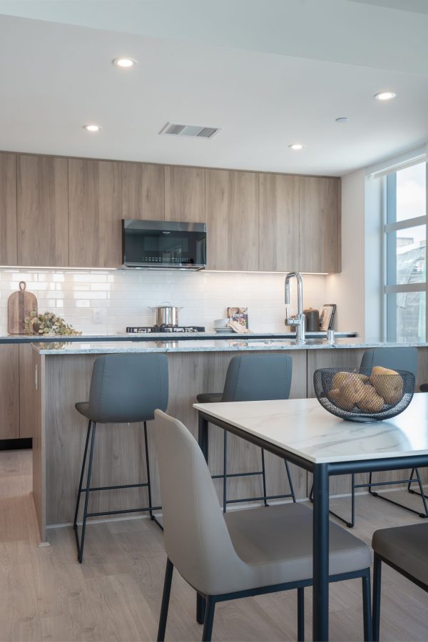 Modern kitchen and dining area with light wood cabinets, gray chairs, a white table, stainless steel appliances, and a black fruit basket with potatoes on the table. Large windows provide natural light.