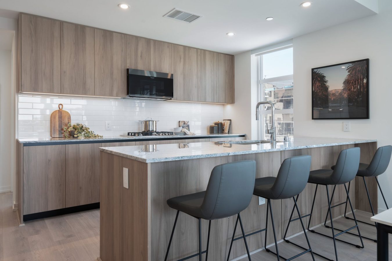 Modern kitchen with light wood cabinets, white tile backsplash, and a marble island with four gray barstools. Stainless steel appliances and a window provide a bright, airy atmosphere. Framed artwork hangs on the wall.