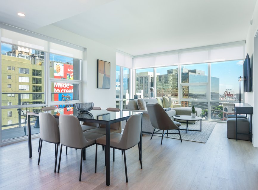 Modern, bright apartment with floor-to-ceiling windows, a dining table with six chairs, lounge chairs, a sofa, TV, and city views featuring colorful buildings and billboards outside.