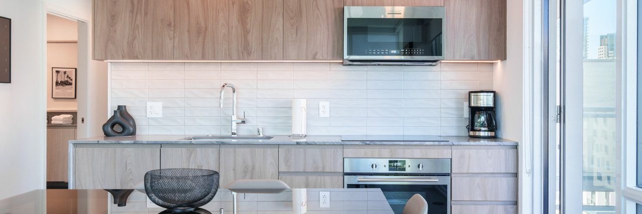 Modern kitchen with light wood cabinets, built-in oven, electric stove, sink, coffee maker, and microwave. A glass dining table with mesh chair is in front. Large windows let in natural light. White tile backsplash.