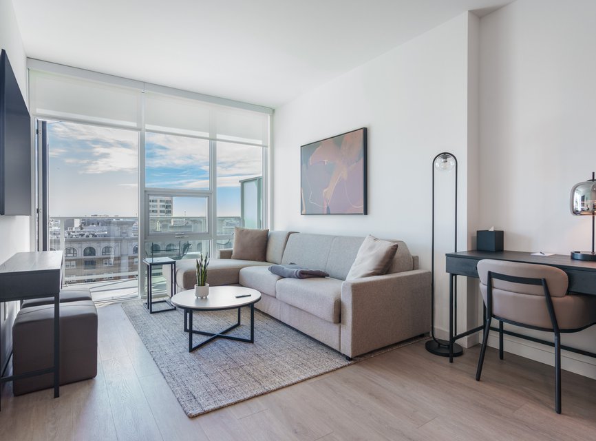 A modern, bright living room with a large window and city view, light gray sofa, round coffee table, rug, and a desk with a chair. Minimalist decor and neutral tones create a clean, airy feel.
