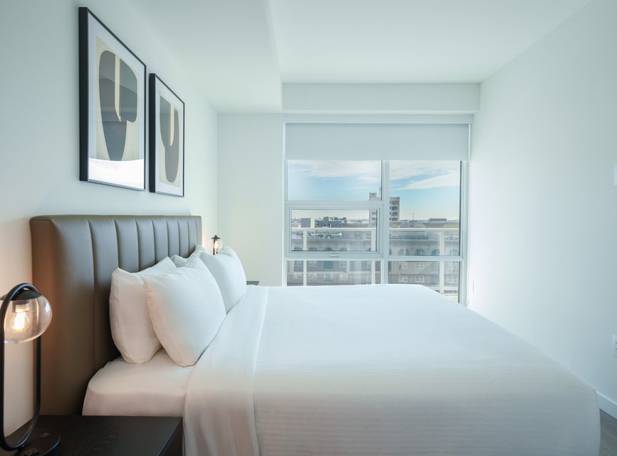 A modern, minimal bedroom with a large bed, white bedding, two abstract art pieces on the wall, a bedside lamp, and a large window with a city view and blue sky outside.