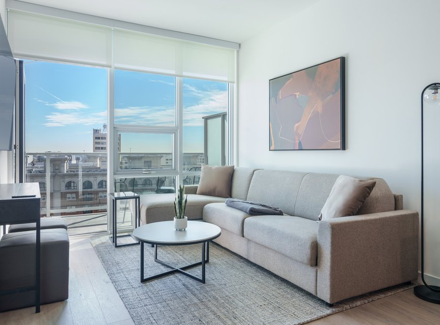 Modern living room with a gray sofa, abstract wall art, round coffee table, floor lamp, and large windows letting in natural light with a cityscape view outside. A small plant sits on the table.