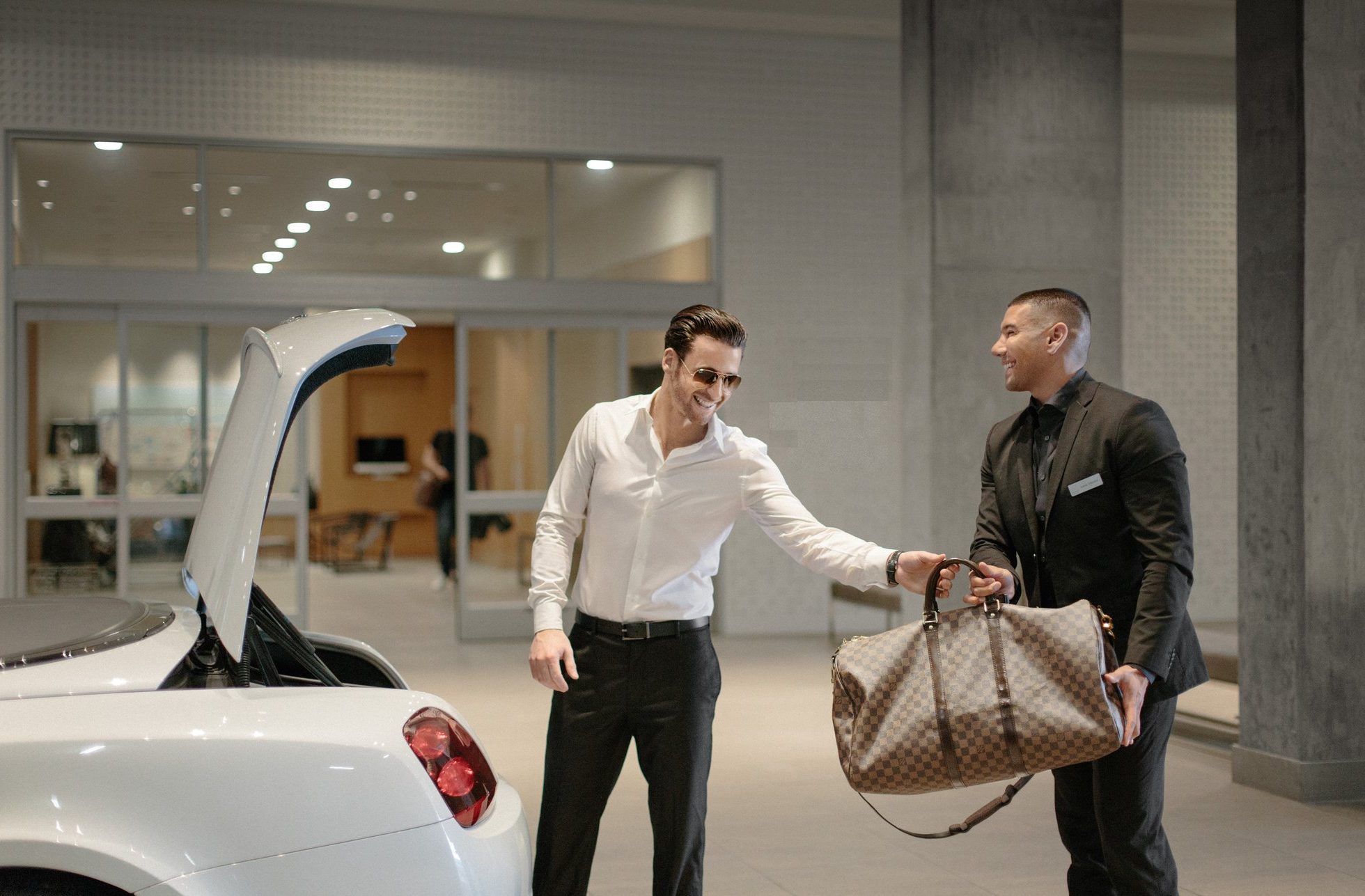 A man in sunglasses hands a large designer duffel bag to a valet standing beside an open car trunk in front of a modern building entrance.