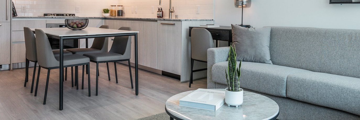 A modern apartment interior featuring a light gray sofa, a small round marble coffee table with books and a potted plant, and a dining area with four chairs beside a minimalist kitchen.