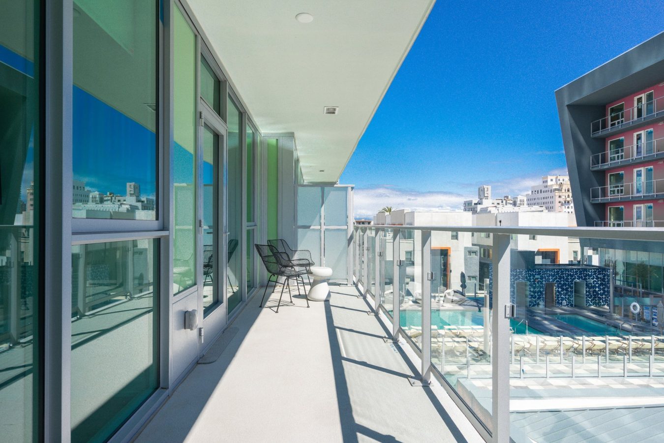 A modern apartment balcony with glass railings, two black chairs, and a small white table overlooks a pool and other contemporary buildings under a bright blue sky.