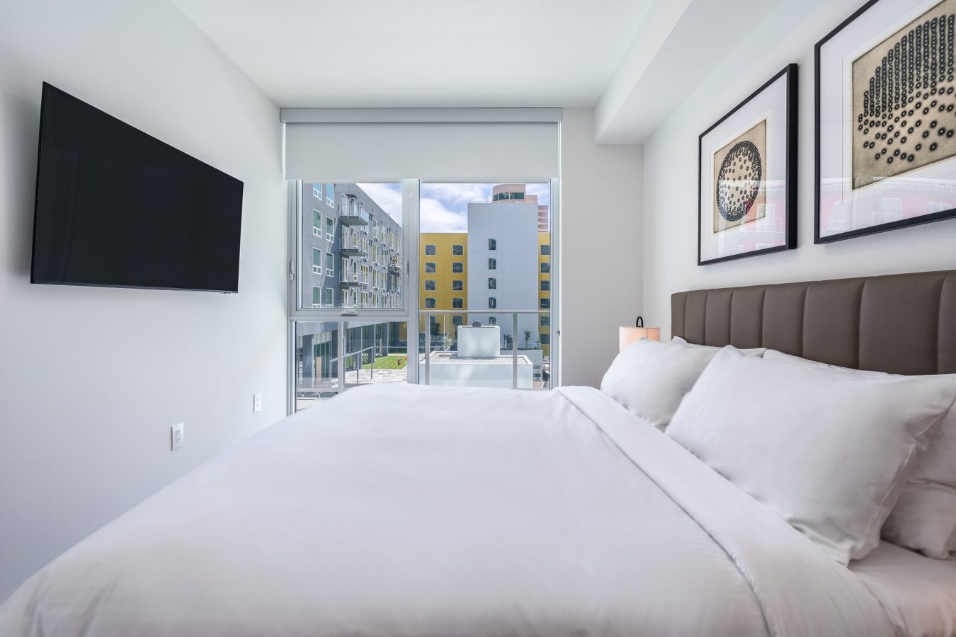 A modern bedroom with a neatly made white bed, wall-mounted TV, framed art, and a large window showing colorful buildings outside. The room is bright and tidy.