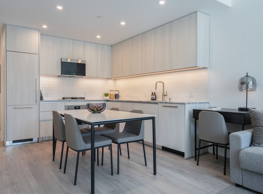 Modern kitchen with light wood cabinets, integrated appliances, and a dining table with four gray chairs. A desk with a chair and lamp sits to the right, next to a gray sofa in a clean, minimalist space.