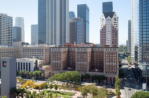 View of downtown Los Angeles with tall modern skyscrapers, historic buildings, and a park with trees and yellow structures in the foreground on a sunny day.