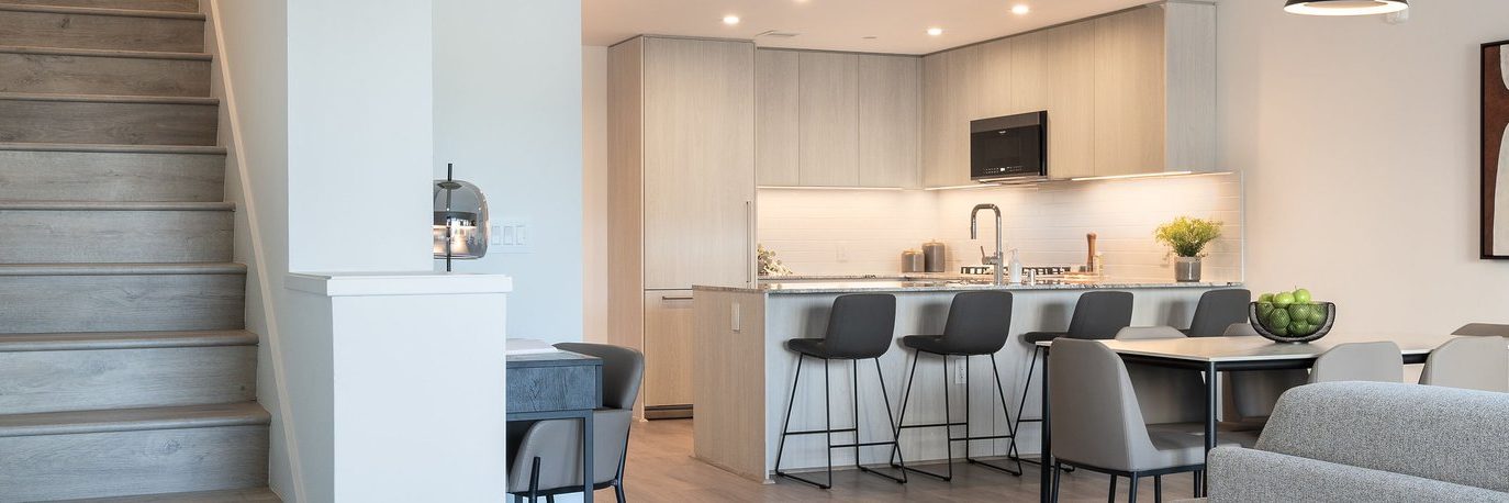 Modern kitchen with light wood cabinets, white backsplash, and a breakfast bar with four black stools. Dining and living areas are visible, along with a staircase on the left side of the image.