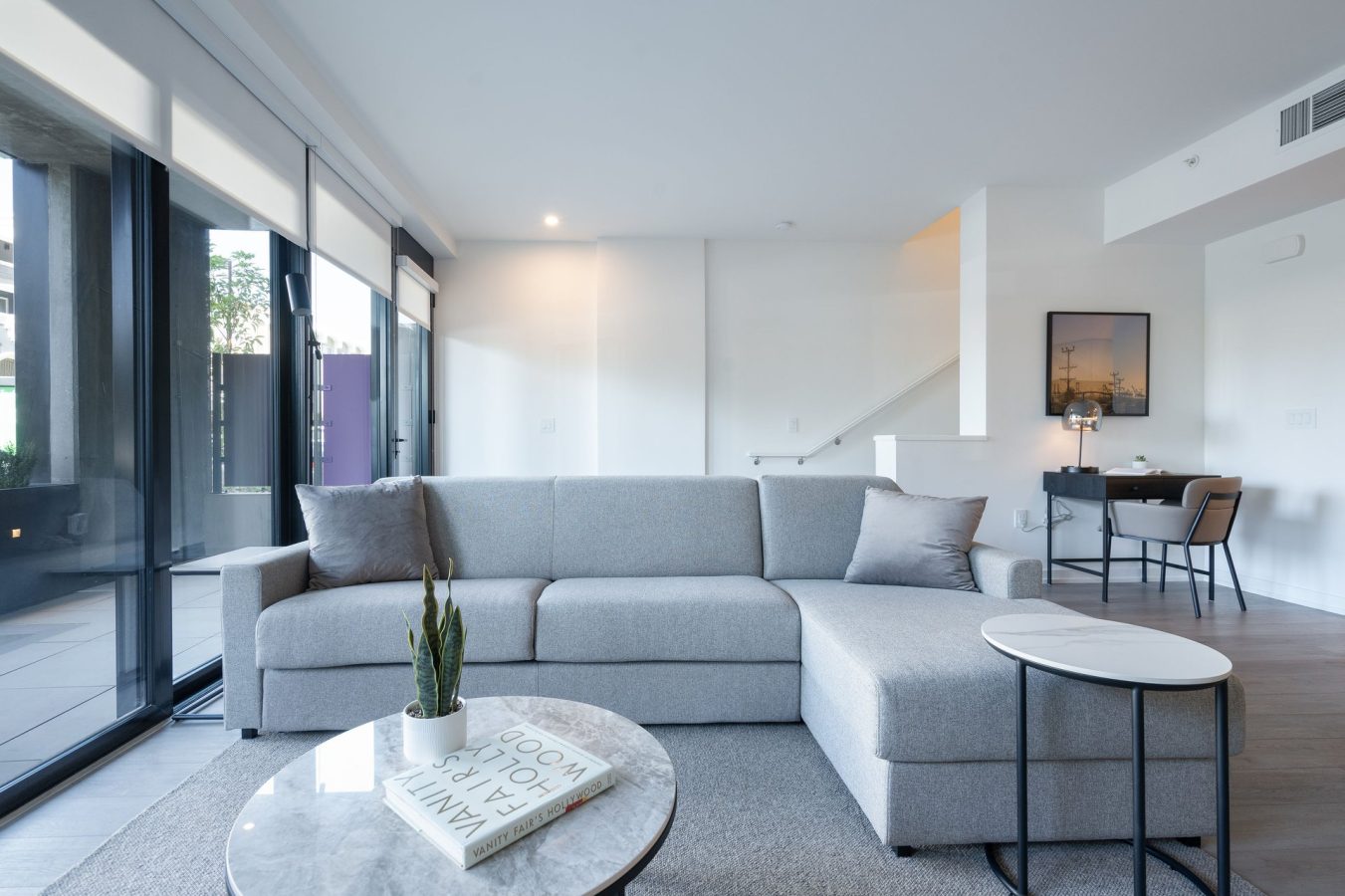 A modern living room with a light gray sectional sofa, round coffee table with a plant and books, large floor-to-ceiling windows, and a small dining area in the background. The space appears bright and minimalistic.