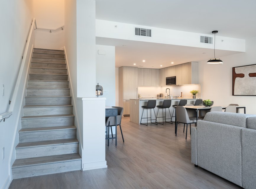 Modern open-concept living space with light wood floors, a staircase on the left, a kitchen with bar seating, dining area, and a gray sofa. Neutral tones and minimalistic decor create a bright, airy atmosphere.