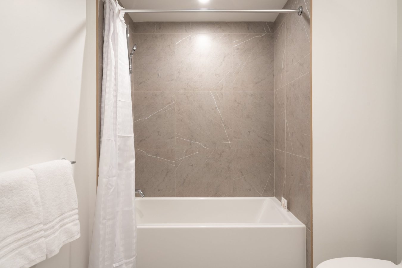 Modern bathroom with a white bathtub, light brown tiled walls, a white shower curtain on a rod, a white towel on a rack, and part of a toilet visible on the right. The space is brightly lit.