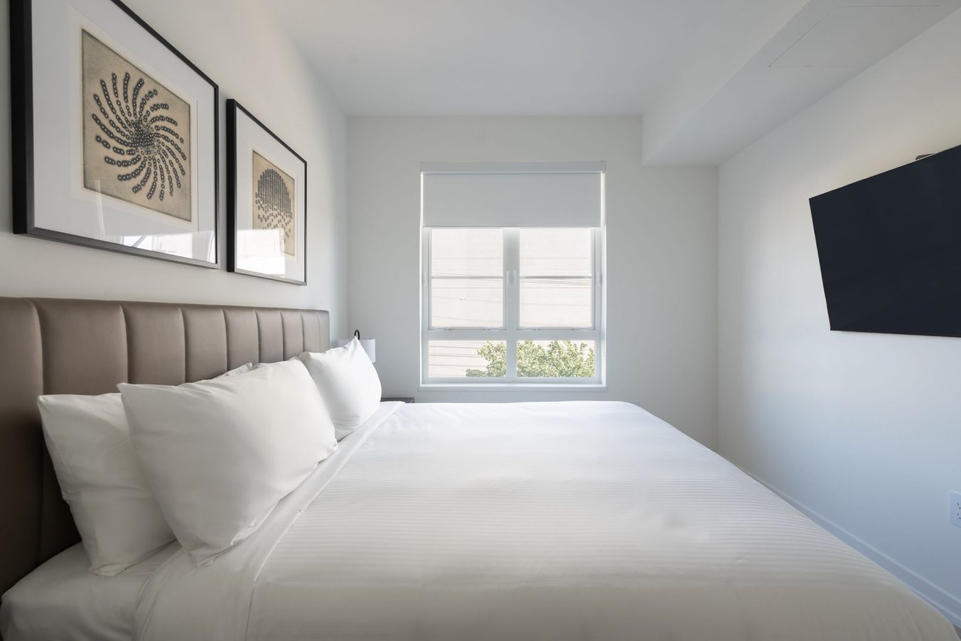 A modern, minimal bedroom with a neatly made bed, white bedding, two framed artworks above the headboard, a window with a roller shade, and a wall-mounted TV on the right.