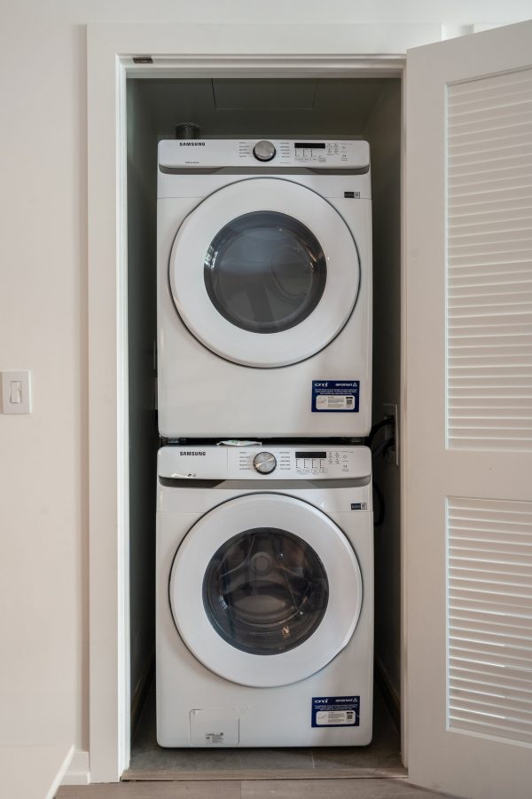 A stacked washer and dryer set in a small, white closet with a partially open louvered door. Both appliances are front-loading and appear clean and modern.