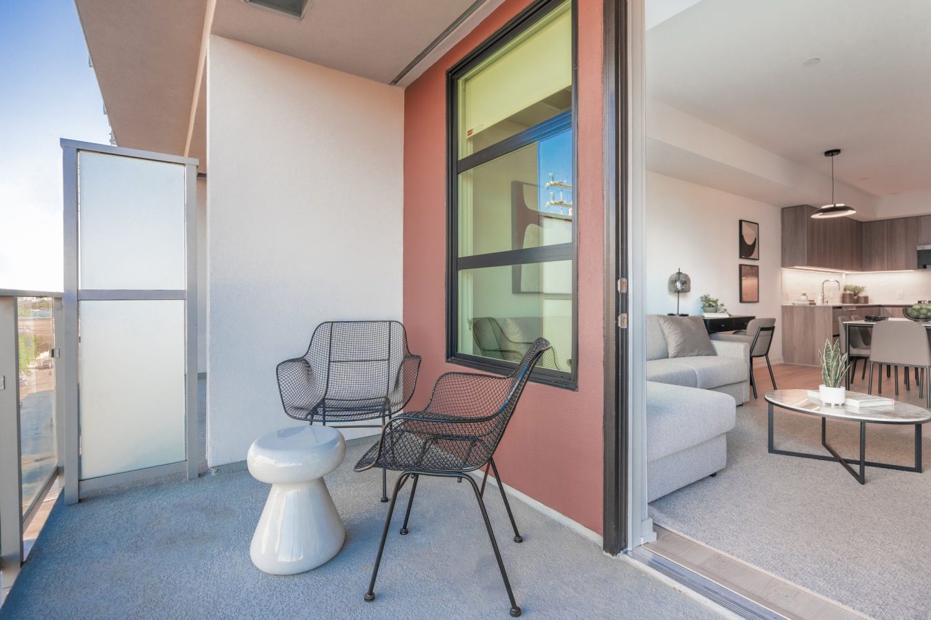 A modern apartment balcony with two black wire chairs and a small white table, adjacent to a living room with a gray sofa and a kitchen in the background. Glass doors separate the balcony from the interior.