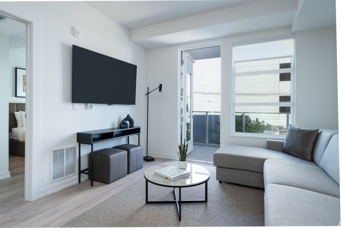 Modern living room with a large wall-mounted TV, a sectional gray sofa, round coffee table, decorative items, and a floor lamp. Large windows and a glass door provide natural light and lead to an outdoor patio.