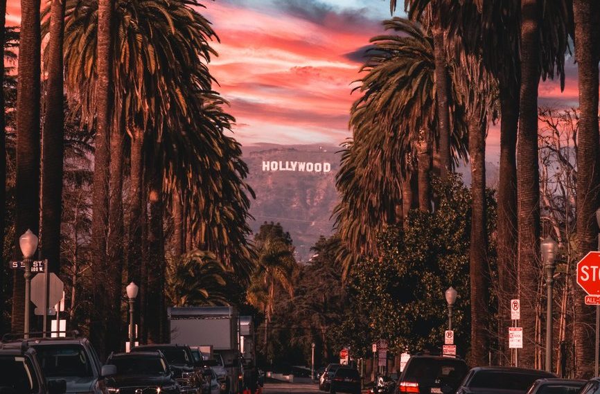 A street lined with tall palm trees leads toward the distant white Hollywood sign on the hills, under a dramatic pink and blue sunset sky, with cars parked along both sides of the road.