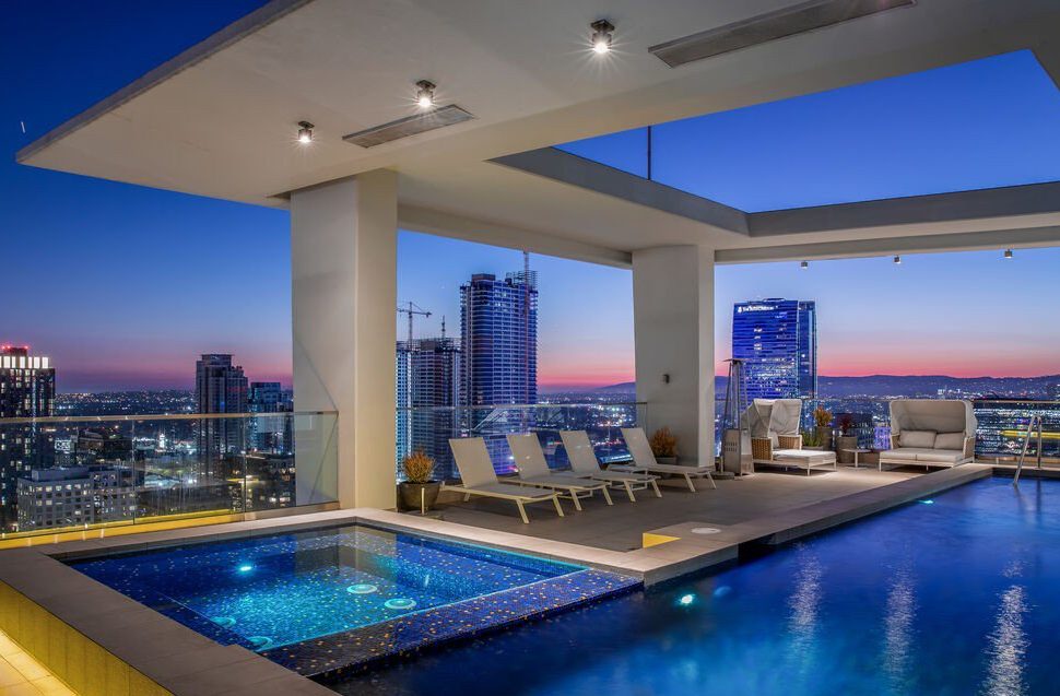 Rooftop pool and lounge area overlooking a city skyline at sunset, with lounge chairs, modern seating, and tall buildings illuminated in the background.