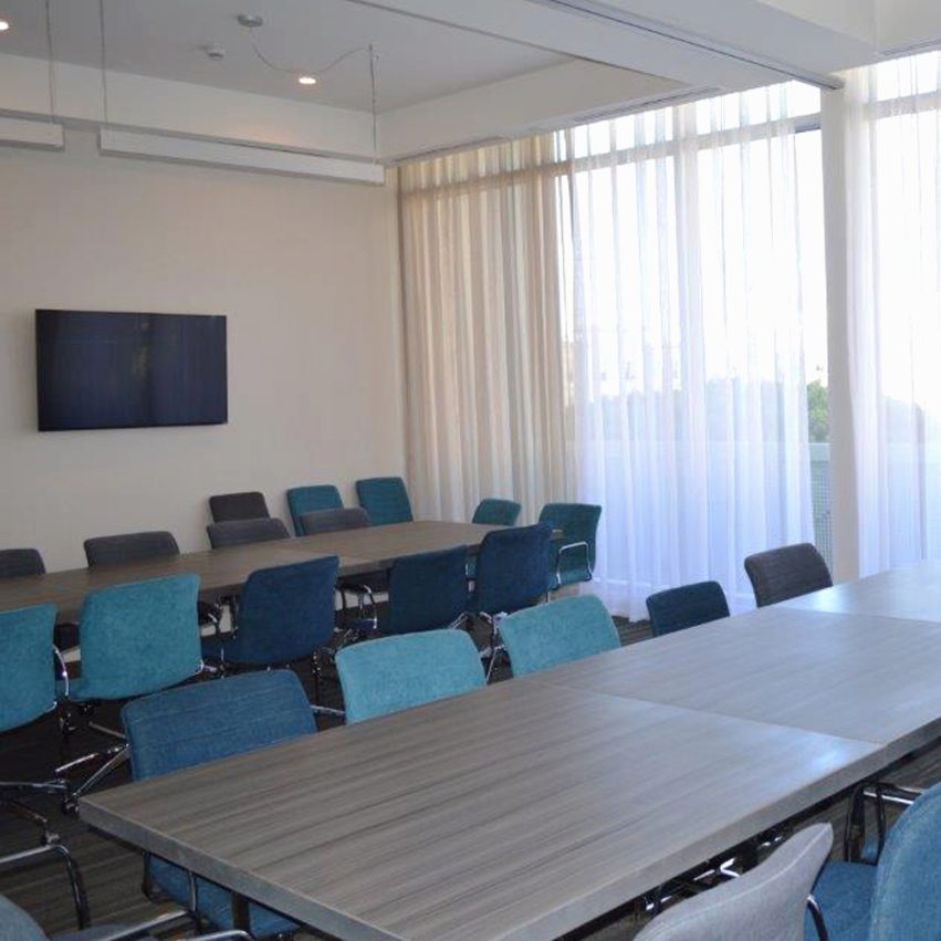 A modern conference room with large windows, sheer curtains, several rectangular tables, and blue and gray office chairs arranged around them. A flat-screen TV is mounted on the wall.