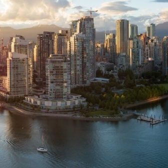 Aerial view of modern high-rise buildings along a waterfront in a city at sunset, with a small sailboat on the water and mountains visible in the background.