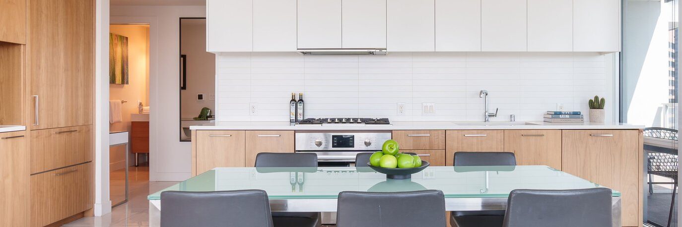 Modern kitchen with light wood cabinets, white countertops, and a glass dining table with black chairs. A bowl of green apples sits on the table; a stove and hood are centered along the back wall. Natural light fills the room.