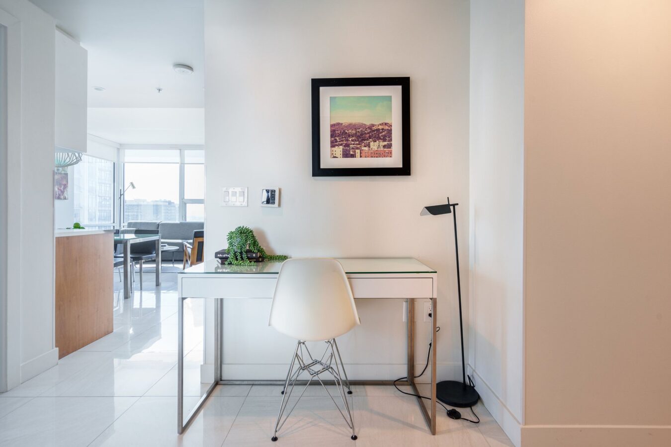A minimalist home office with a white desk and chair, a small potted plant, a black floor lamp, and a framed picture on the wall, set in a bright, modern apartment.