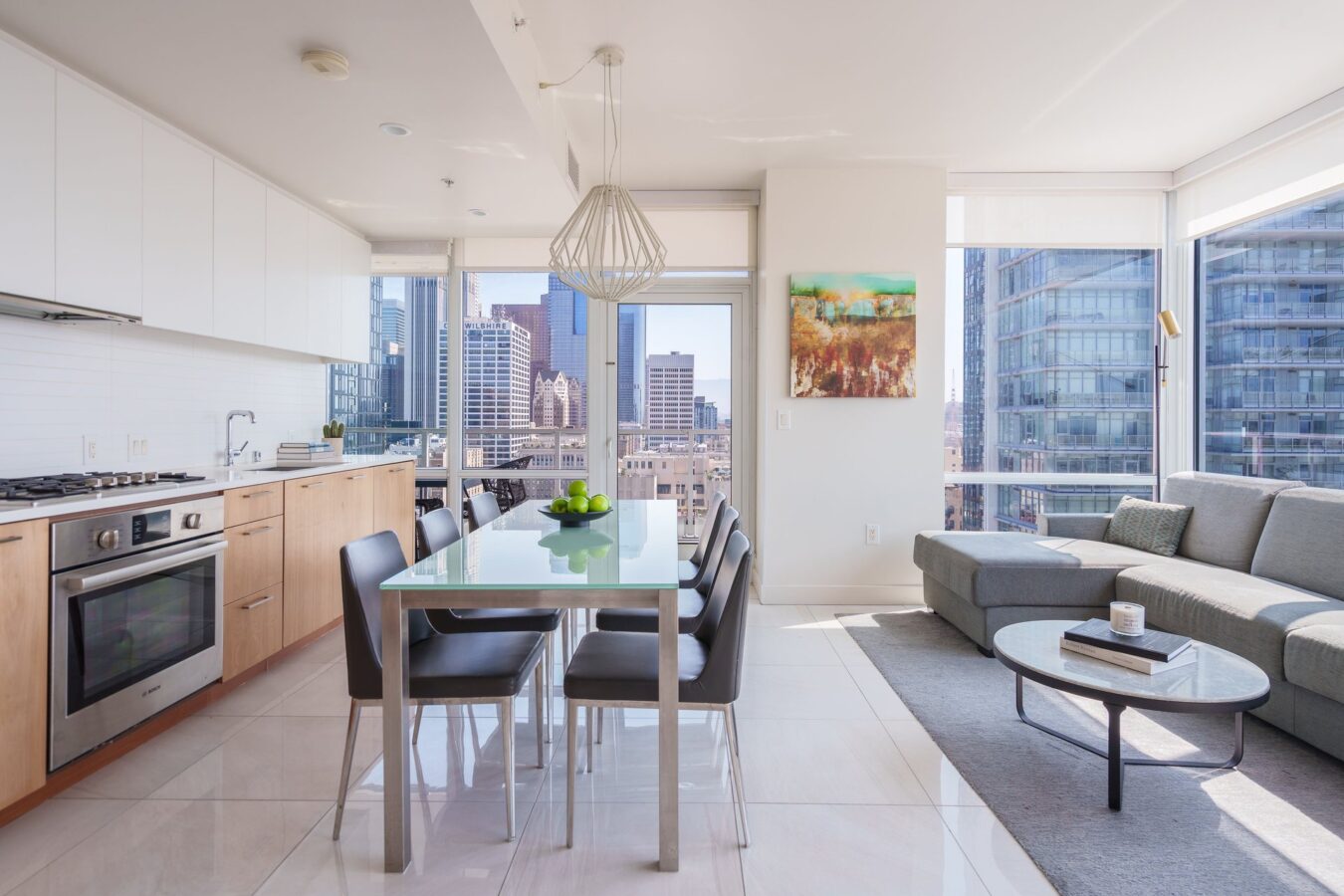 A modern, bright apartment with a kitchen, dining table, and living area. Large windows offer a city view. The space features light wood cabinets, a gray sofa, a glass dining table, and wall art.