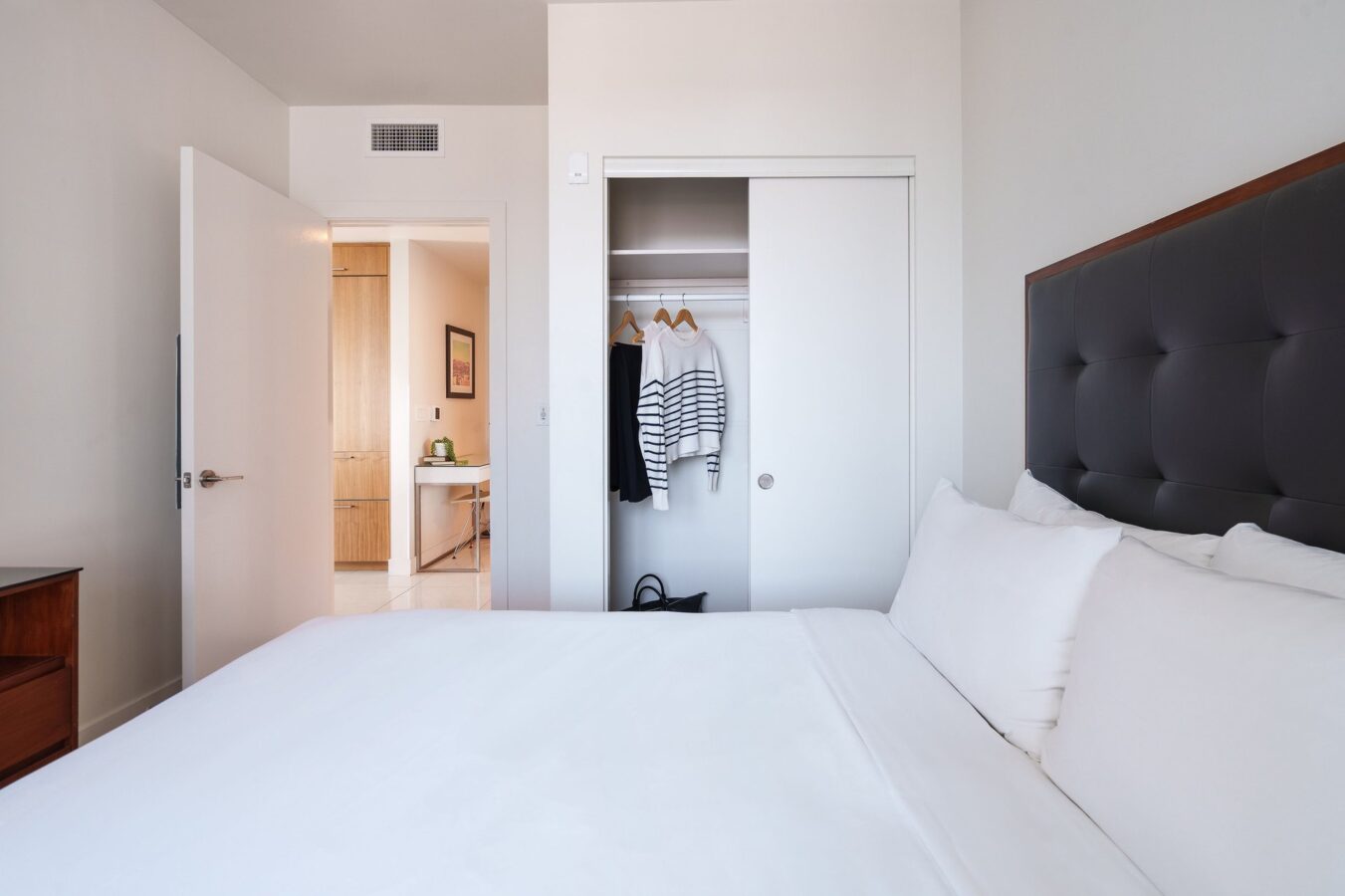 A tidy, modern bedroom with a neatly made bed, a dark headboard, an open closet with hanging clothes, and an open door revealing a glimpse of a hallway or another room.