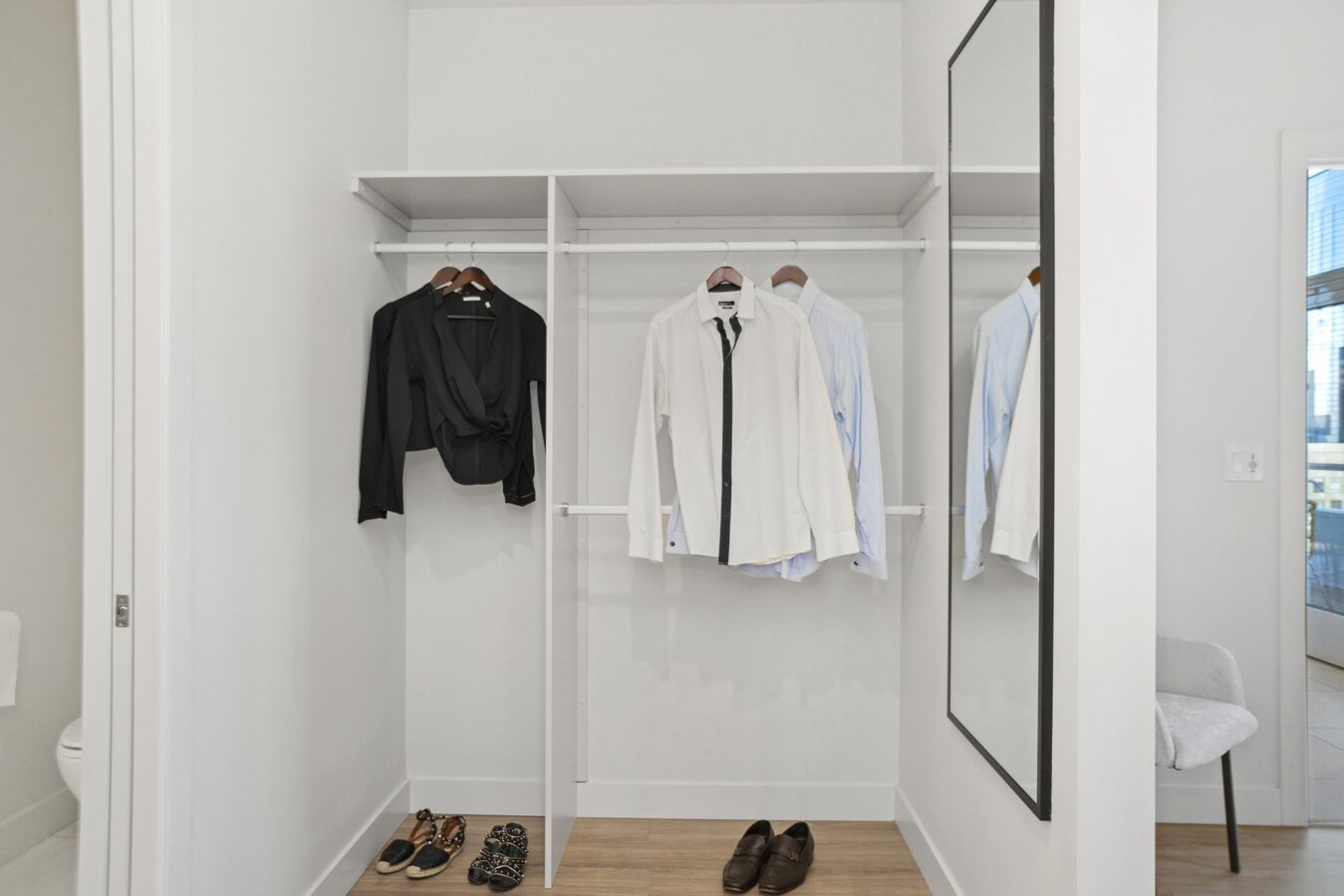 Open closet with hanging shirts and a blouse, dress shoes, and sandals on the floor. A large wall mirror and a chair are visible; the space has light walls and wood flooring.