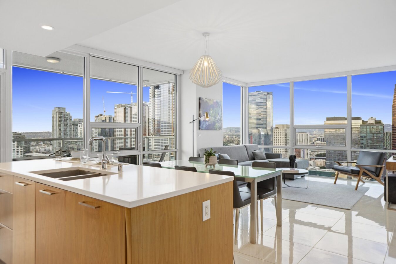 Modern high-rise apartment with large windows, city skyline views, an open-plan kitchen with a white island, wood cabinets, a dining table, and a living area with a gray sofa and a TV on the wall.