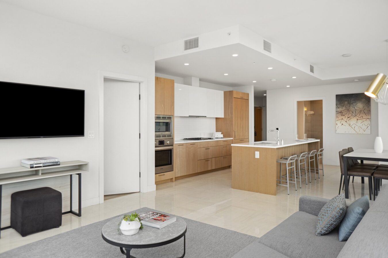 Modern open-plan living space with a kitchen featuring light wood cabinets, a breakfast bar with stools, a gray sofa, a wall-mounted TV, and a dining area with contemporary decor in neutral tones.