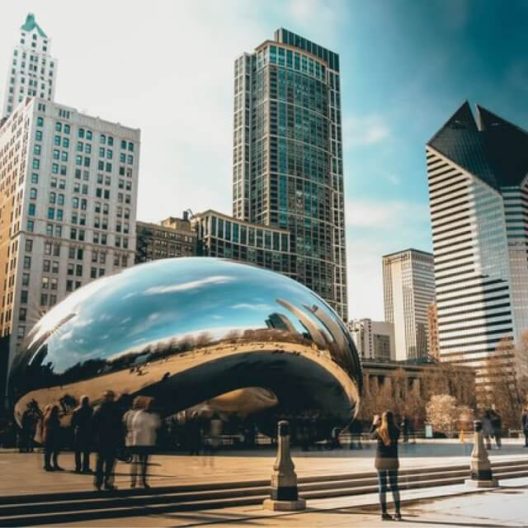 People gather around the reflective Cloud Gate sculpture, also known as The Bean, in Chicago’s Millennium Park, with tall buildings and a partly cloudy sky in the background.