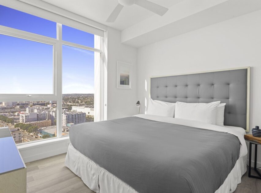 Modern bedroom with a large bed featuring a gray headboard and gray bedding, light wood floor, a nightstand, and a window offering a city view under a clear blue sky.