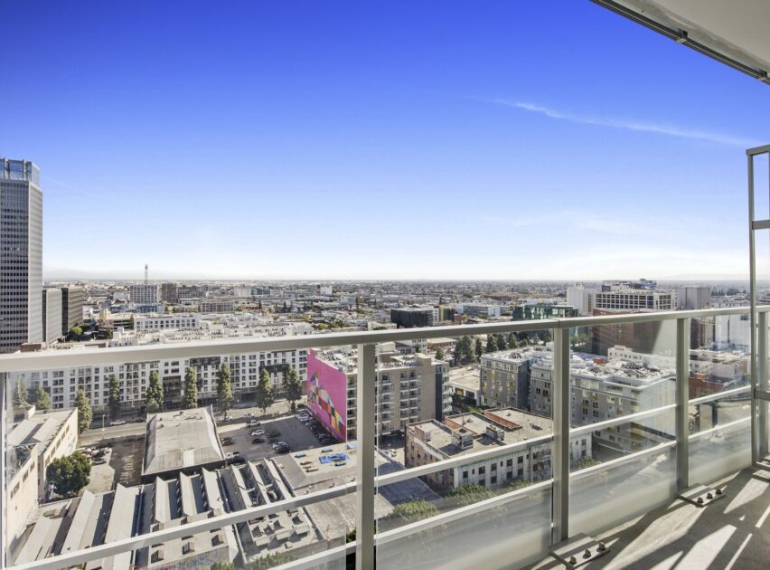 View from a high-rise balcony overlooking a cityscape with modern buildings, a colorful mural, clear blue sky, and sunlight casting shadows on the glass railing and balcony floor.