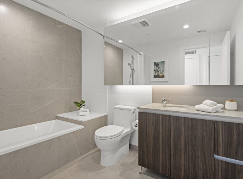 Modern bathroom with beige tile walls, a bathtub, white toilet, wooden vanity with sink, large mirror, folded towels, a small plant, and framed artwork on the wall. Bright, clean, and minimalist design.