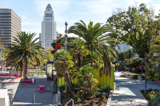 A sunny outdoor plaza with palm trees, pink tables and chairs, and modern buildings in the background, including a tall white tower with a pointed top. Lush greenery surrounds the area.