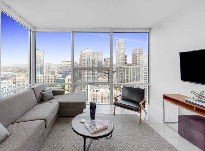 Modern living room with large windows showing a city skyline view, gray sectional sofa, black chair, round coffee table with books, and wall-mounted TV on a wooden console.
