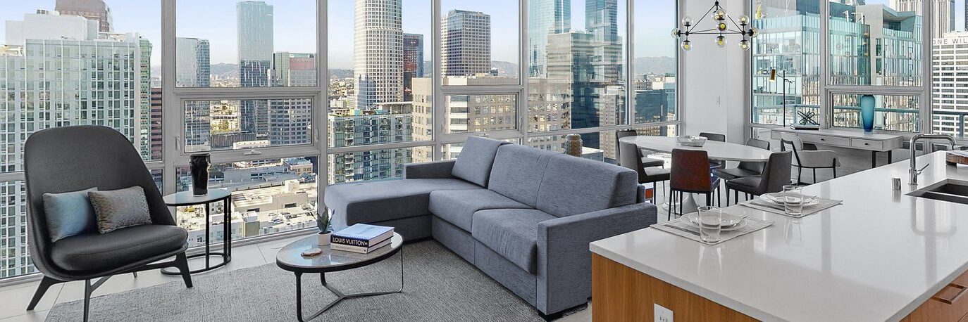 Modern apartment with large windows showing a city skyline, featuring a gray sofa, armchair, round coffee table, dining table with chairs, and a kitchen island with place settings in an open-plan living space.