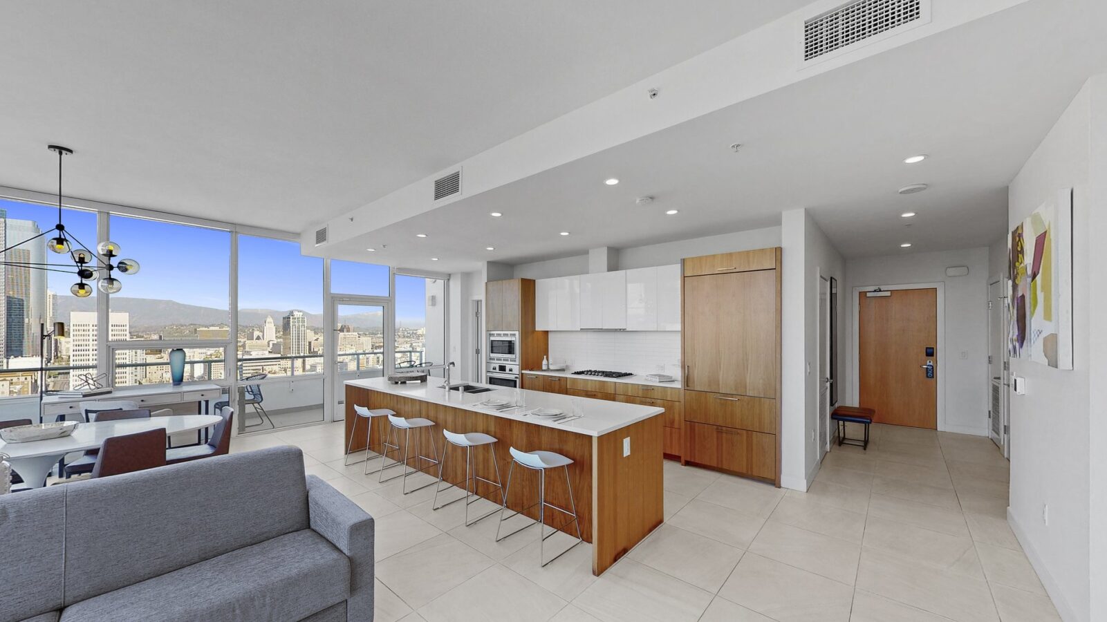 Modern open-plan kitchen and dining area in a high-rise apartment, featuring sleek wood cabinetry, a white island with barstools, large windows with city views, and neutral-colored tile flooring.