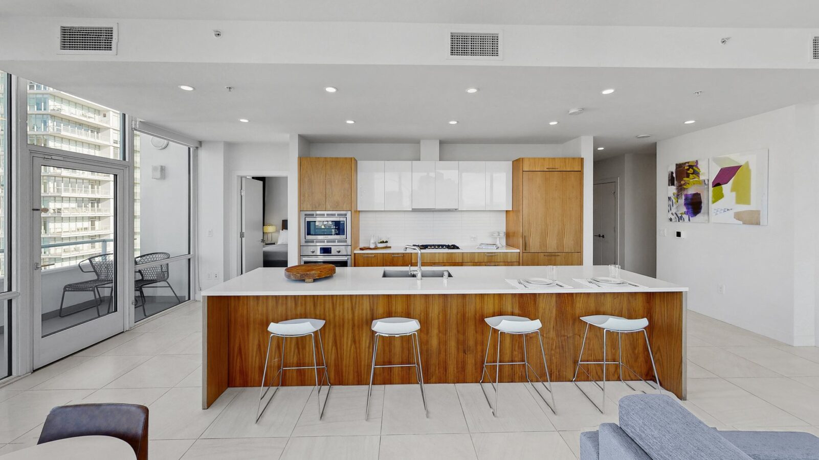 Modern kitchen with a large wooden island, four white barstools, built-in appliances, white cabinets, and a balcony with glass doors; abstract artwork hangs on the wall to the right.