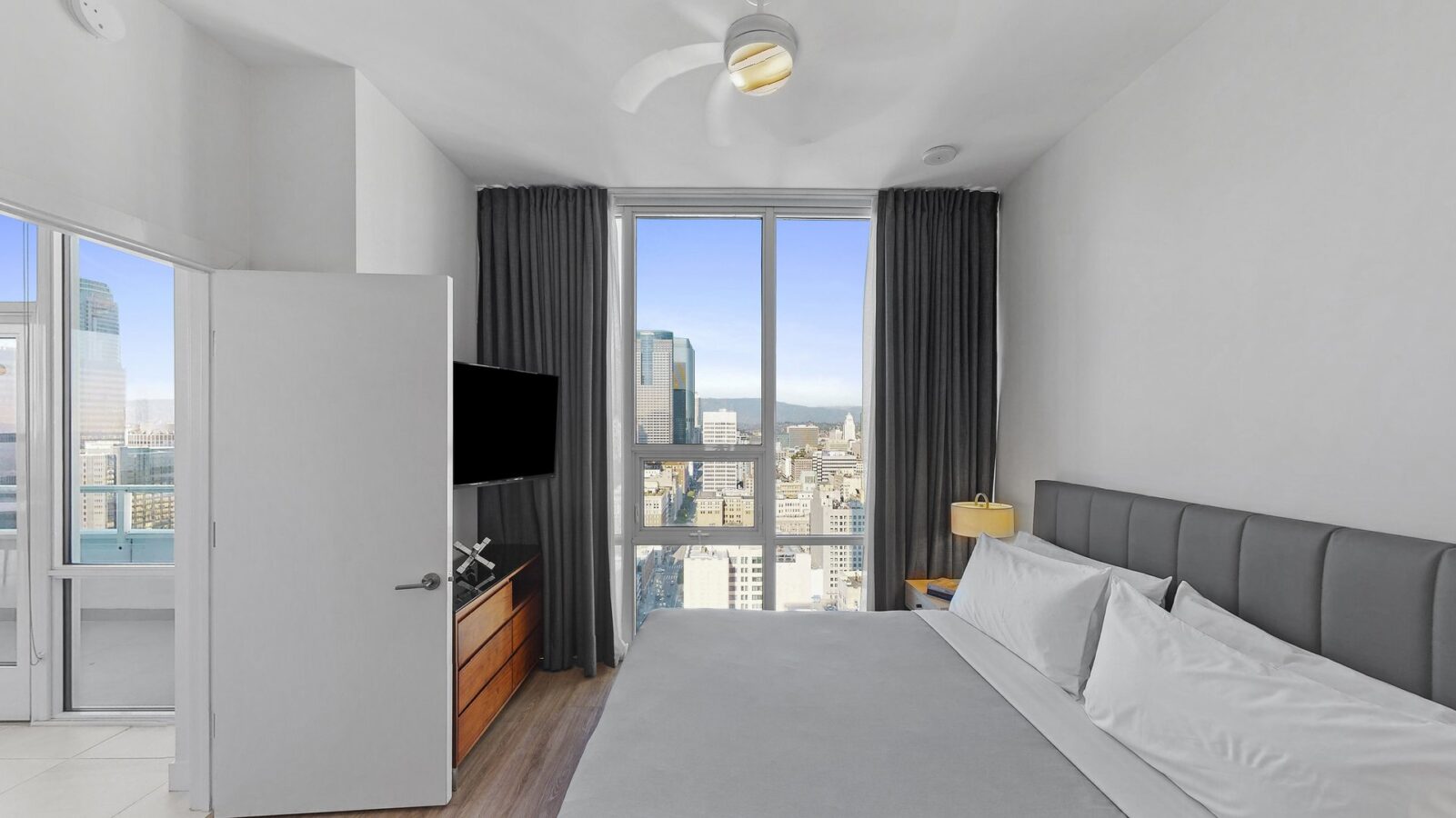 A modern bedroom with a large bed, gray headboard, TV on a wooden dresser, and floor-to-ceiling window offering a city skyline view. Dark curtains frame the window, and a ceiling fan hangs above.