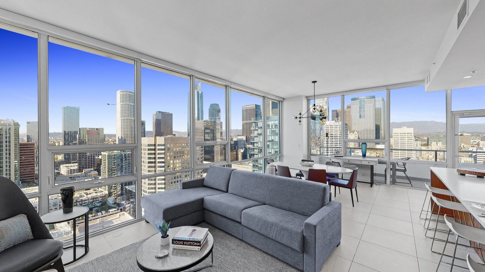 Modern apartment living room with large windows offering a panoramic view of a city skyline. The room has a gray sofa, coffee table, dining area, and kitchen with bar stools, all in bright, natural light.