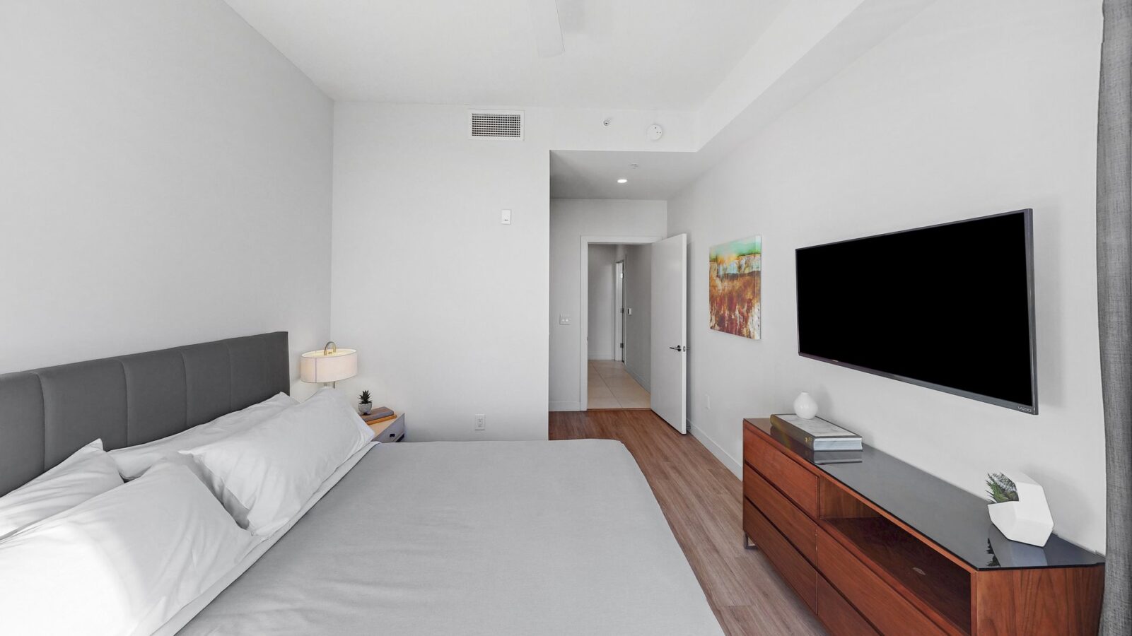 A modern, minimalist bedroom with a neatly made bed, gray headboard, wooden dresser, wall-mounted TV, a bedside lamp, and a landscape painting on a white wall. The door is open to a bright hallway.