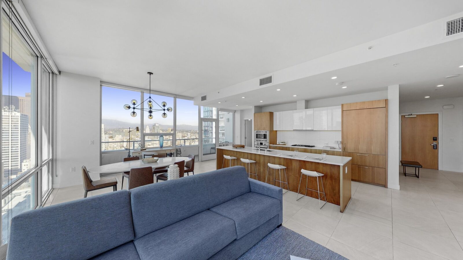 Modern open-concept apartment with large windows, city views, a blue sofa, dining area, and sleek kitchen with wooden cabinets, white countertops, and bar stools. Bright, minimalist decor throughout the space.