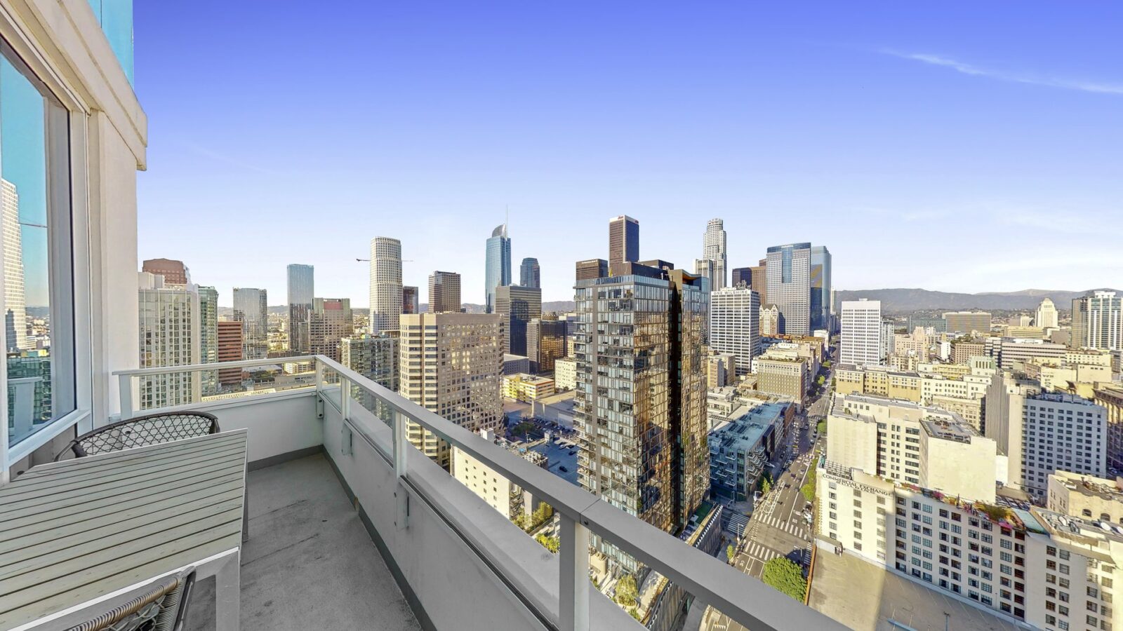 A spacious balcony with a table and two chairs overlooks a city skyline filled with tall buildings under a clear blue sky.