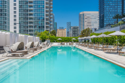 A modern outdoor swimming pool surrounded by lounge chairs, cabanas, and umbrellas, set amid tall city buildings under a clear blue sky.