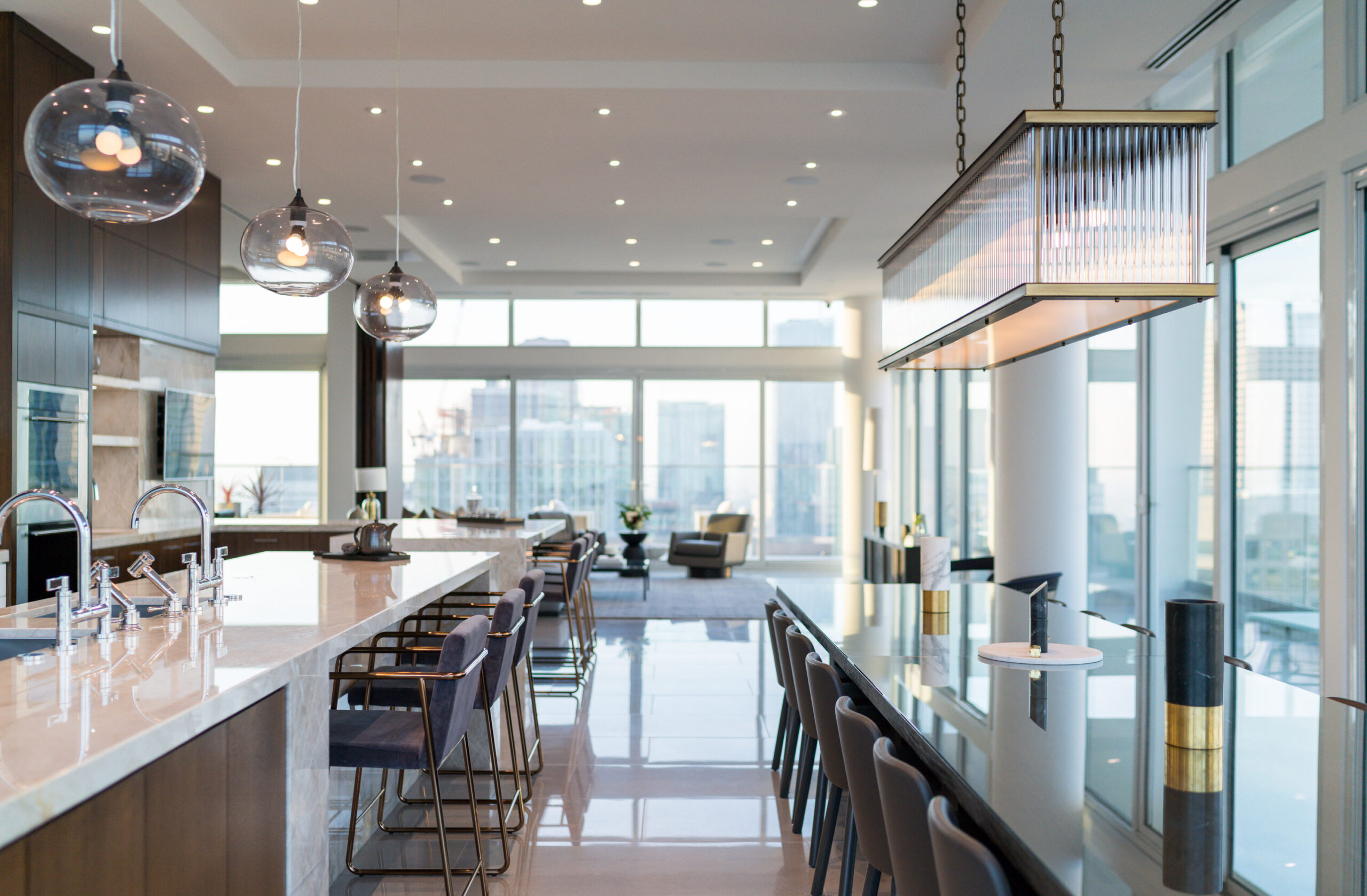 Modern, open-concept kitchen and dining area with large windows, marble countertops, stylish pendant lights, bar stools, and a cityscape view in the background.